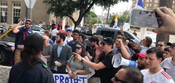 Protestors gather outside a bar in Texas with one person shouting into a megaphone while another person holds a sign reading 'Protect TX kids'