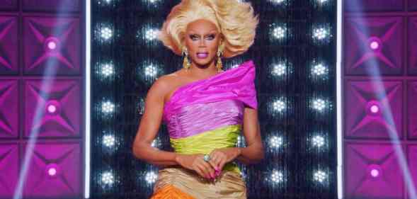 RuPaul wears a one should dress in pink, yellow, tan and orange with a blonde wig on the set of Drag Race All Stars 7