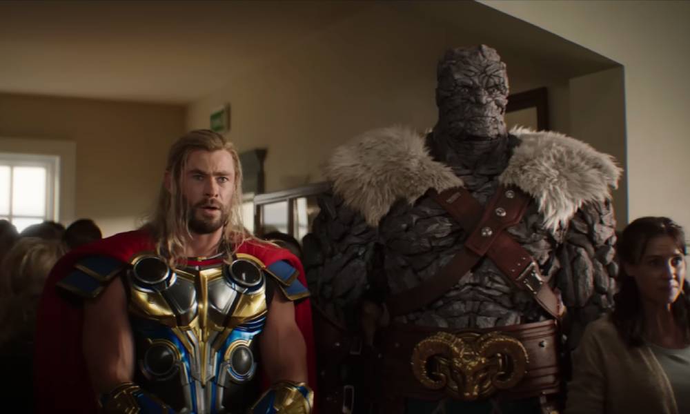 Chris Hemsworth plays Thor, wearing a gold and blue coloured armoured chest plate with a red close. He is standing next to Korg (Taika Waititi) who is a tall alien covered in what appears to be rock-like skin. Korg has two bands crossing his chest and fuzzy shoulder pads