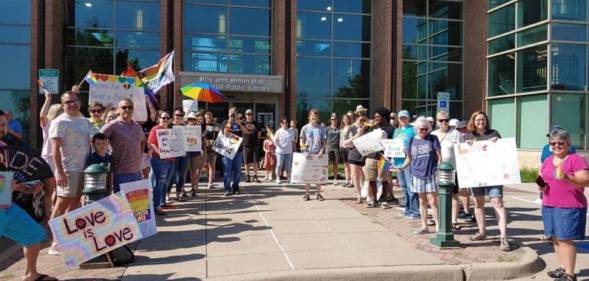 A crowd of people holding up signs in support of the LGBTQ+ community, Pride flags and colourful umbrellas gather outside the Roy and Helen Hall Library in Texas