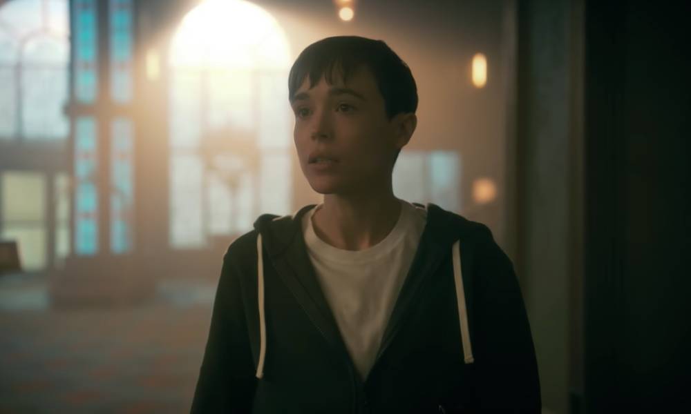 Elliot Page appears as his character from the Netflix show The Umbrella Academy. The character, Viktor, is wearing a white t-shirt and blue hooded sweatshirt as he stands indoors and speaks to someone off screen