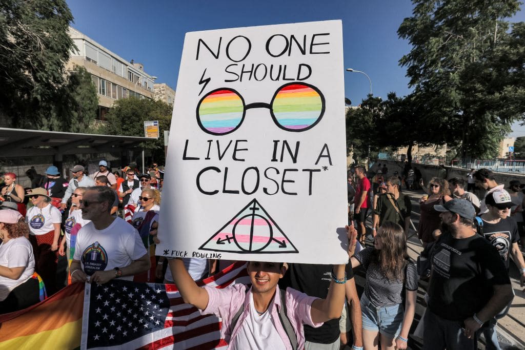 A Pride participant marches with a sign reading in English "no one should live in a closet", with round, Harry-Potter style glasses with a rainbow motif and the deathly hallows symbol (also from Harry potter) in the colour of the trans pride flag.