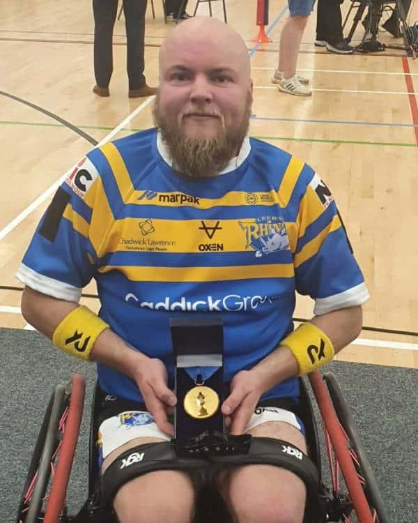 Verity Smith wears a yellow and blue striped rugby jersey while seated in a wheelchair. He is holding a gold medal in a black box on his lap