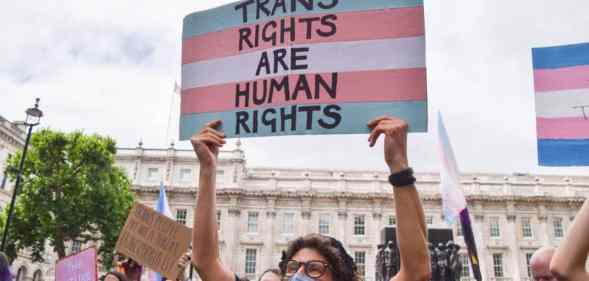 A person wearing a face mask designed in the colours of the trans Pride flag holds up a sign with the colours of the trans Pride flag that reads "Trans rights are human rights" above their head during a protest