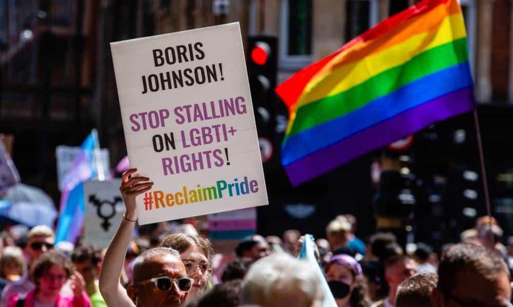 A person attending London Pride holds up a white sign reading "Boris Johnson! Stop stalling on LGBTI+ rights #ReclaimPride" with a rainbow LGBTQ+ flag seen in the background