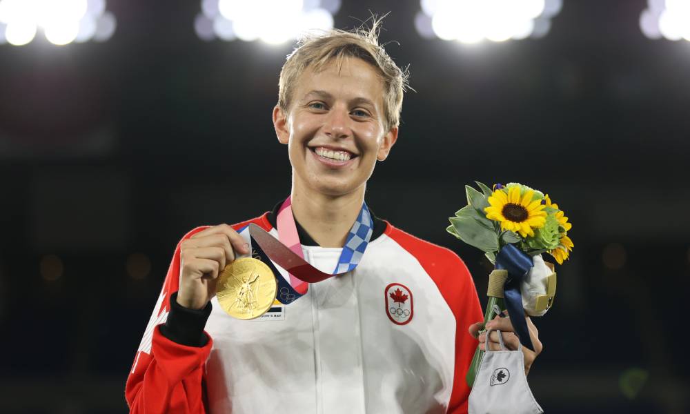 Team Canada professional footballer Quinn holds up their Olympic gold medal and a small group of flowers as they wear a red and white jacket