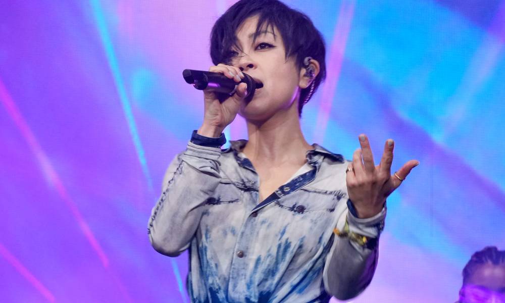 Hikaru Utada wears a blue top as she sings in front of people gathered for Coachella. They are holding up a microphone as they perform in front of a purple and blue background