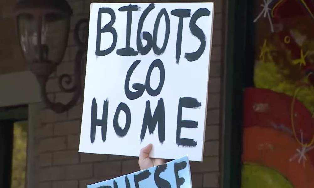 A person holds up a white sign reading 'Bigots go home' in black writing. The person is part of a crowd of counter-protestors outside a drag event in Houston, Texas