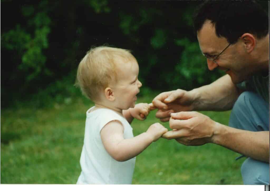 Alice as a baby. Her father holds her hands as he helps her take her first steps on a green lawm