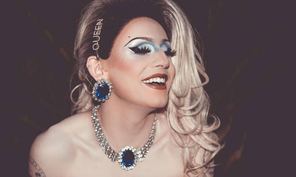 Lin, a drag queen, wears a long-haired blond wig with a hair pin reading 'queen' in rhinestones. She is also wearing a bejewelled necklace and matching earrings