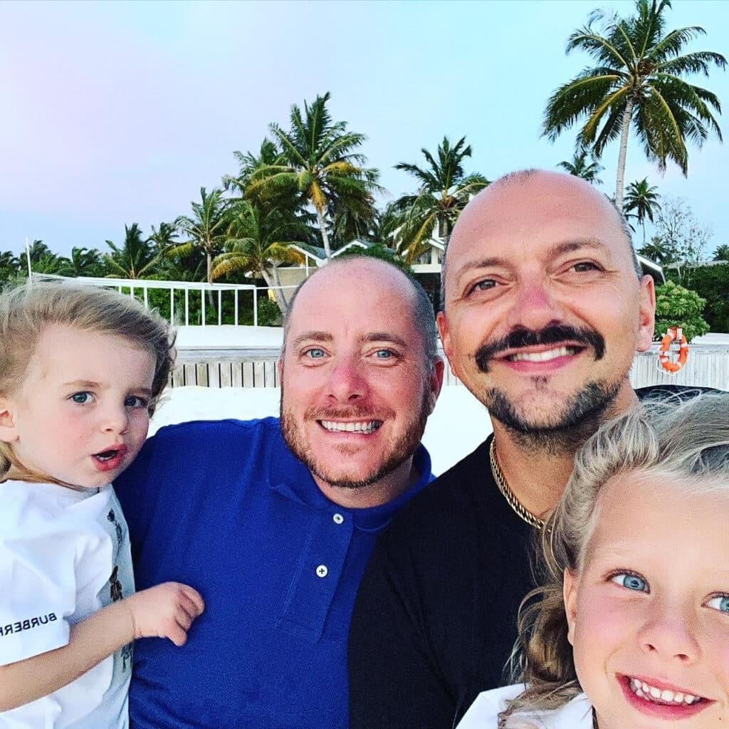 Michael and Wes on holiday with their kids. They're smiling at the camera, with palm trees visible in the background.