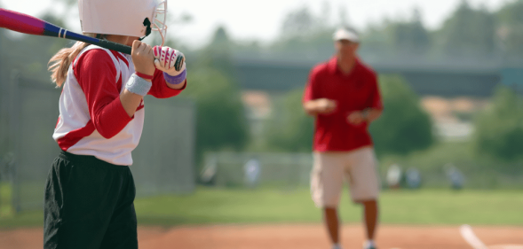 Stock image of a young girl holding a bat while a man coaches her in the background