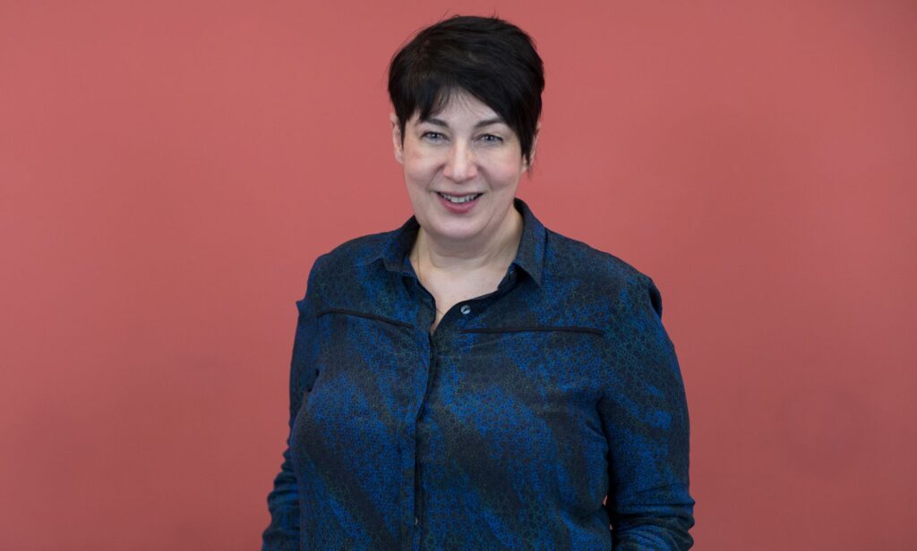 Joanne Harris wears a blue shirt as she poses in front of a pink backdrop