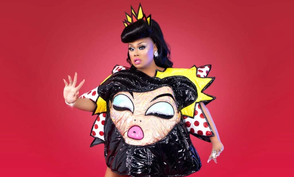 In this photograph, drag queen Jiggly Caliente wears a black wig with a pop art themed outfit in red and yellow colours. The outfit has a drawing of her face on it. She is standing in front of a red background with one hand posed upwards