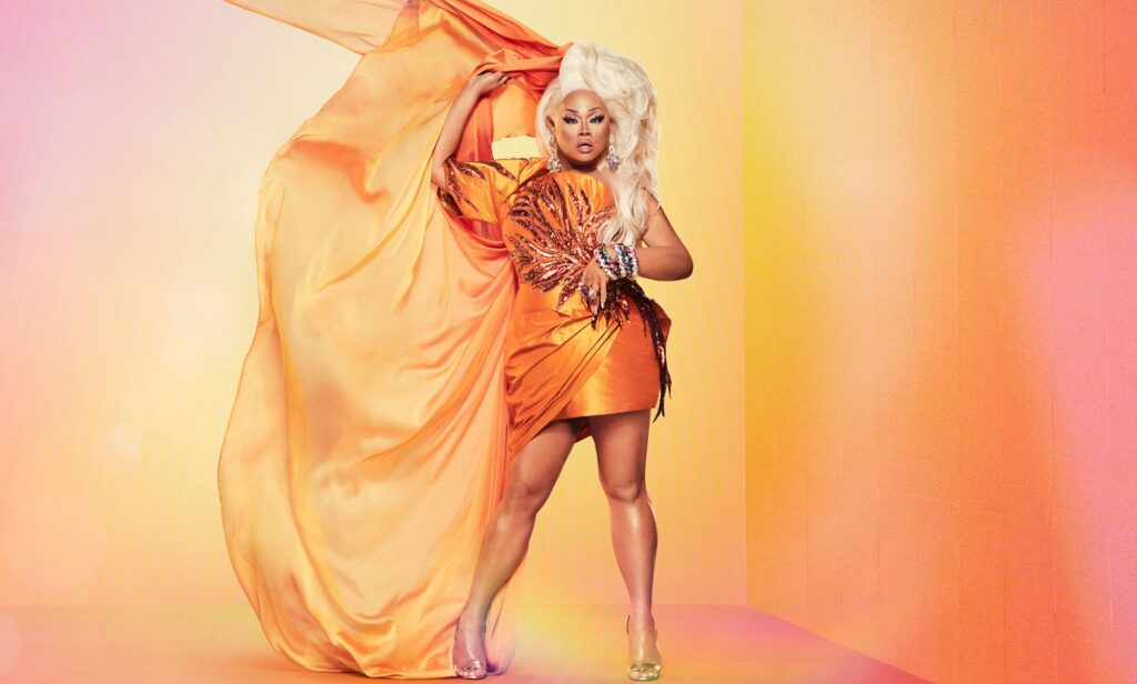 In this photograph, Drag Race Philippines host Jiggly Caliente wears a short orange dress with posing with a matching piece of orange fabric, which is held in her hands, waving next to her. She is wearing a blonde wig as she stands in front of an orange, peach, yellow and pink background