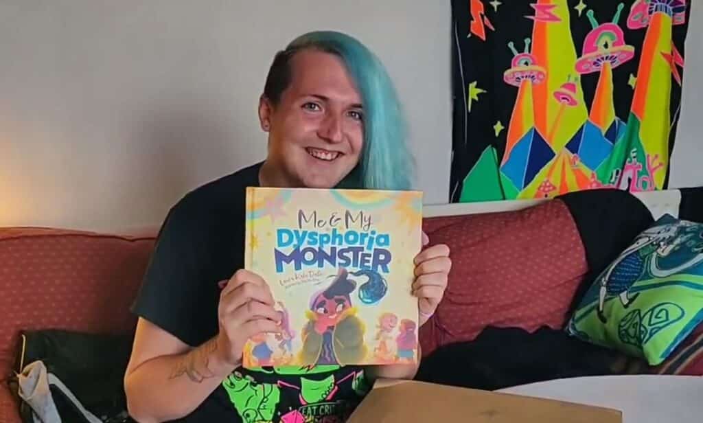 In this photograph, a woman with blue hair holds up a copy of her illustrated children's book 'Me and My Dysphoria Monster', which focuses on the life of a young trans girl. 