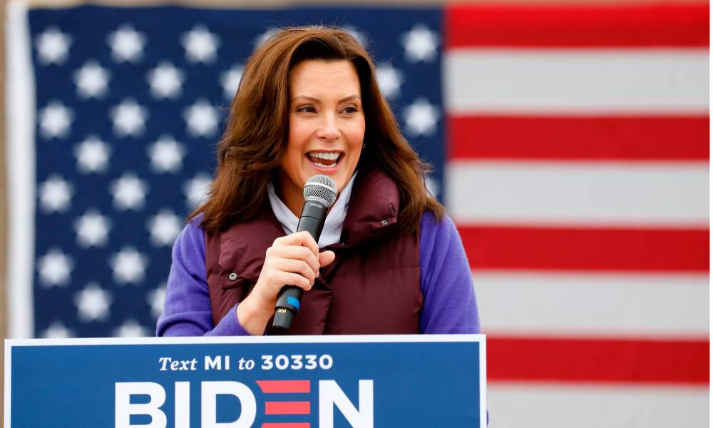 Michigan Governor Gretchen Whitmer holds a microphone as she stands in front of an American flag and at a podium with a sign for Joe Biden's presidential race. She is wearing a purple outfit