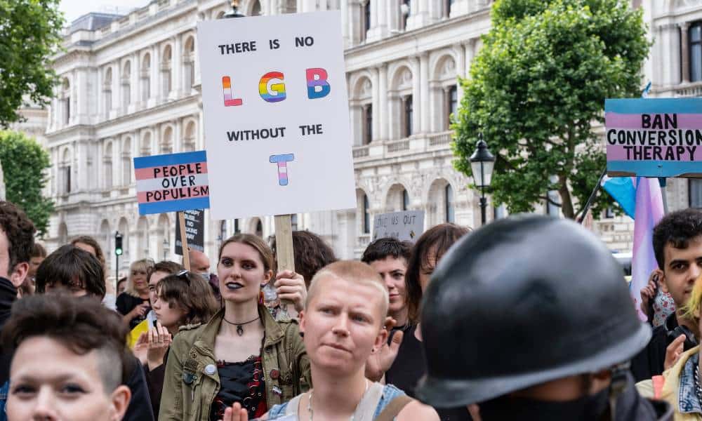 People gather in a crowd to call on the UK government to put forward legislation protecting all LGBTQ+ people from conversion therapy. One person holds up a sign that reads 'There is no LGB without the T'
