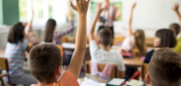 A stock photo of children with their hands raised in a classroom, facing a teacher and a whiteboard