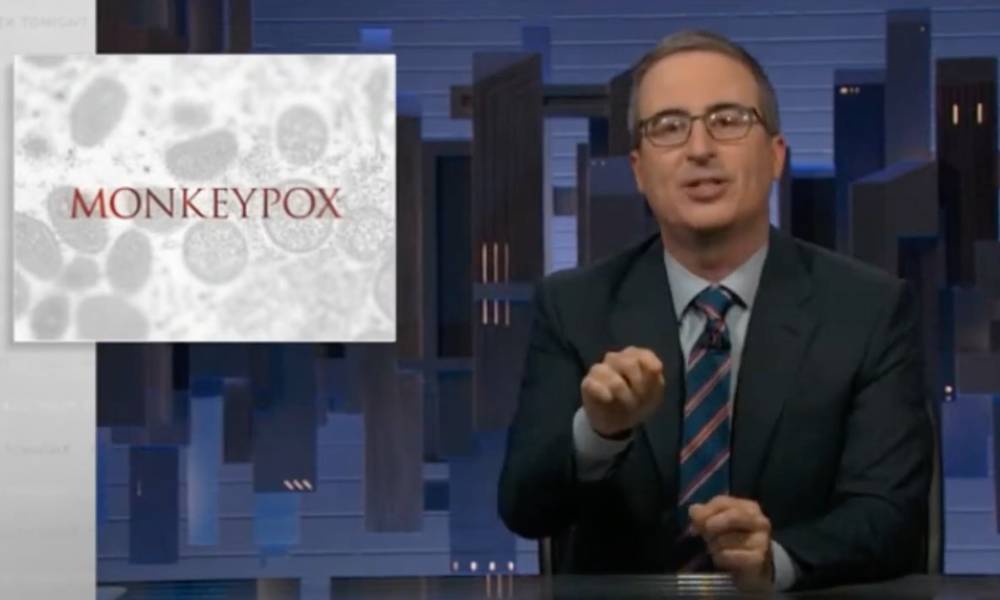 John Oliver wears a blue shirt, striped tie and dark suit jacket on the set of HBO Max's Last Week Tonight. There is an graphic depicting monkeypox in the corner