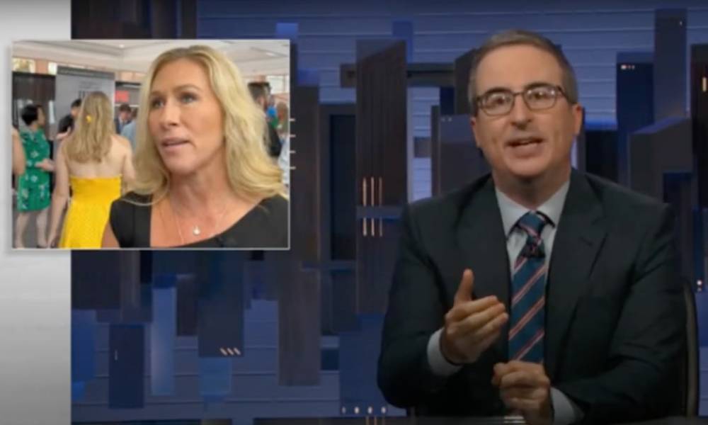 John Oliver wears a blue shirt, striped tie and dark suit jacket on the set of HBO Max's Last Week Tonight. There is an image of Georgia congresswoman Marjorie Taylor Greene in the corner wearing a dark top as she speaks to someone off-camera
