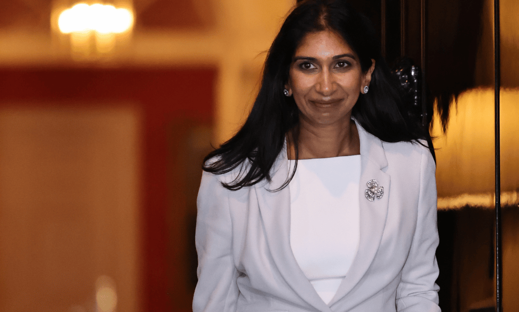 Suella Braverman wears a white and grey outfit as she walks out a doorway