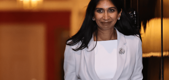 Suella Braverman wears a white and grey outfit as she walks out a doorway