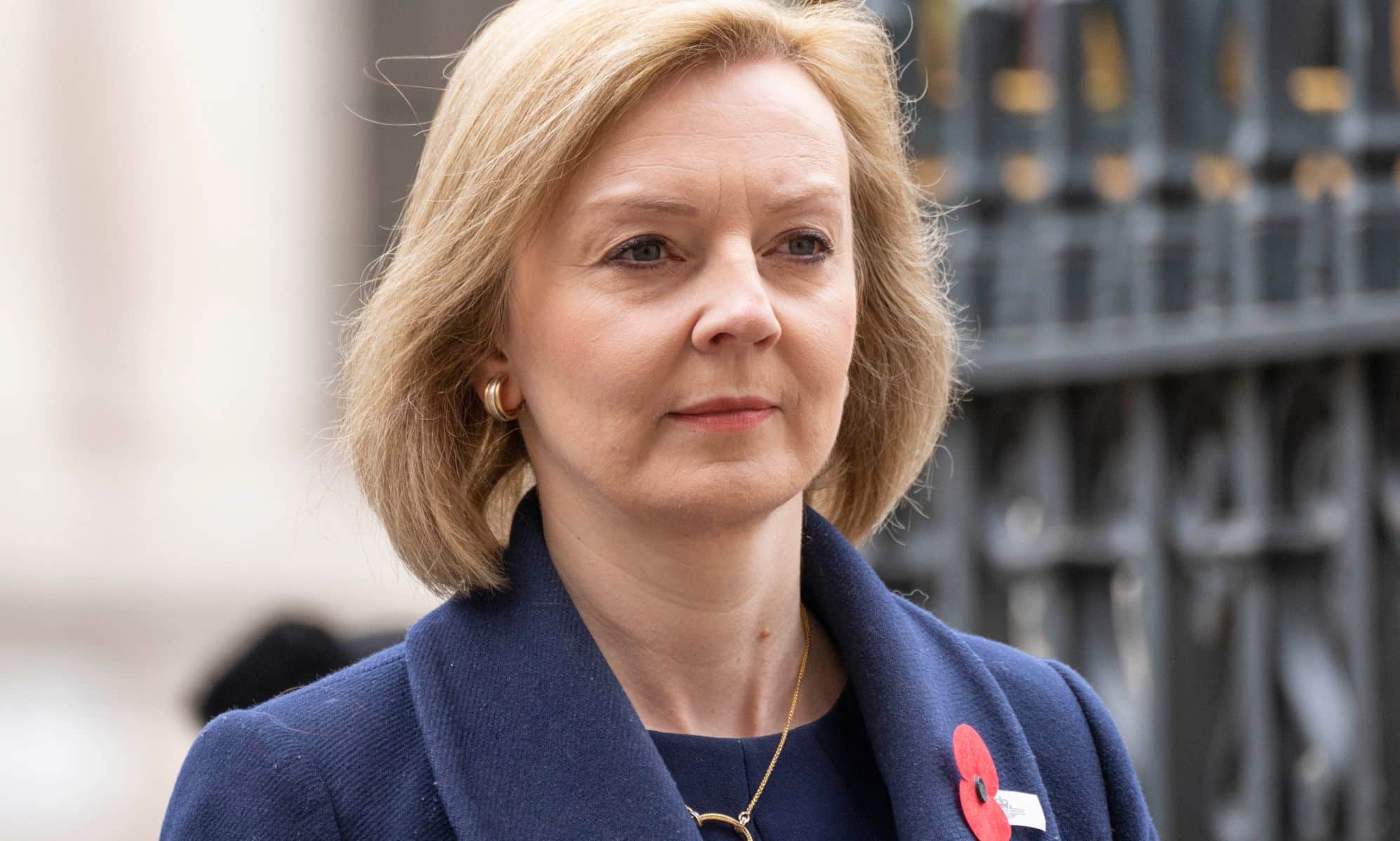 Liz Truss wears a dark blue jacket with a red poppy as she stands outside