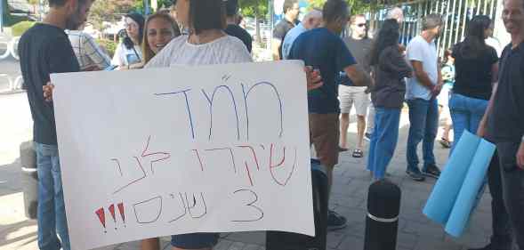 A parent stands outside the demonstration area holding a sign in hebrew.