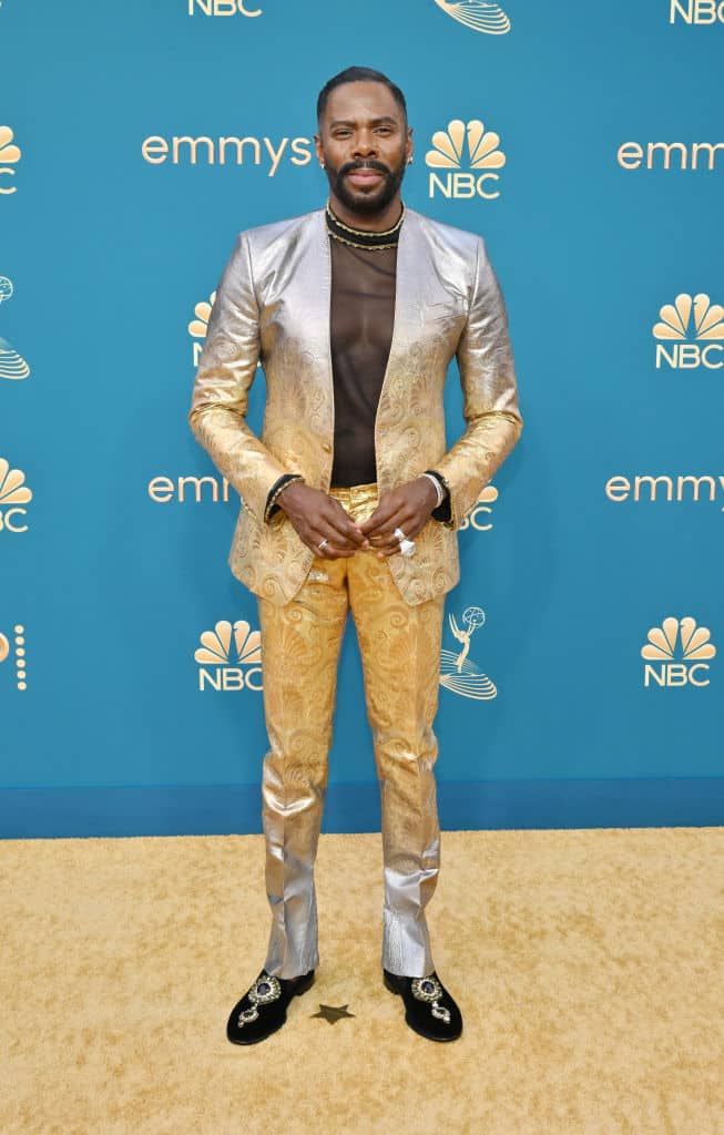 Colman in a silver jacquard suit with a sheer crew neck top, standing on a gold carpet against a blue backdrop