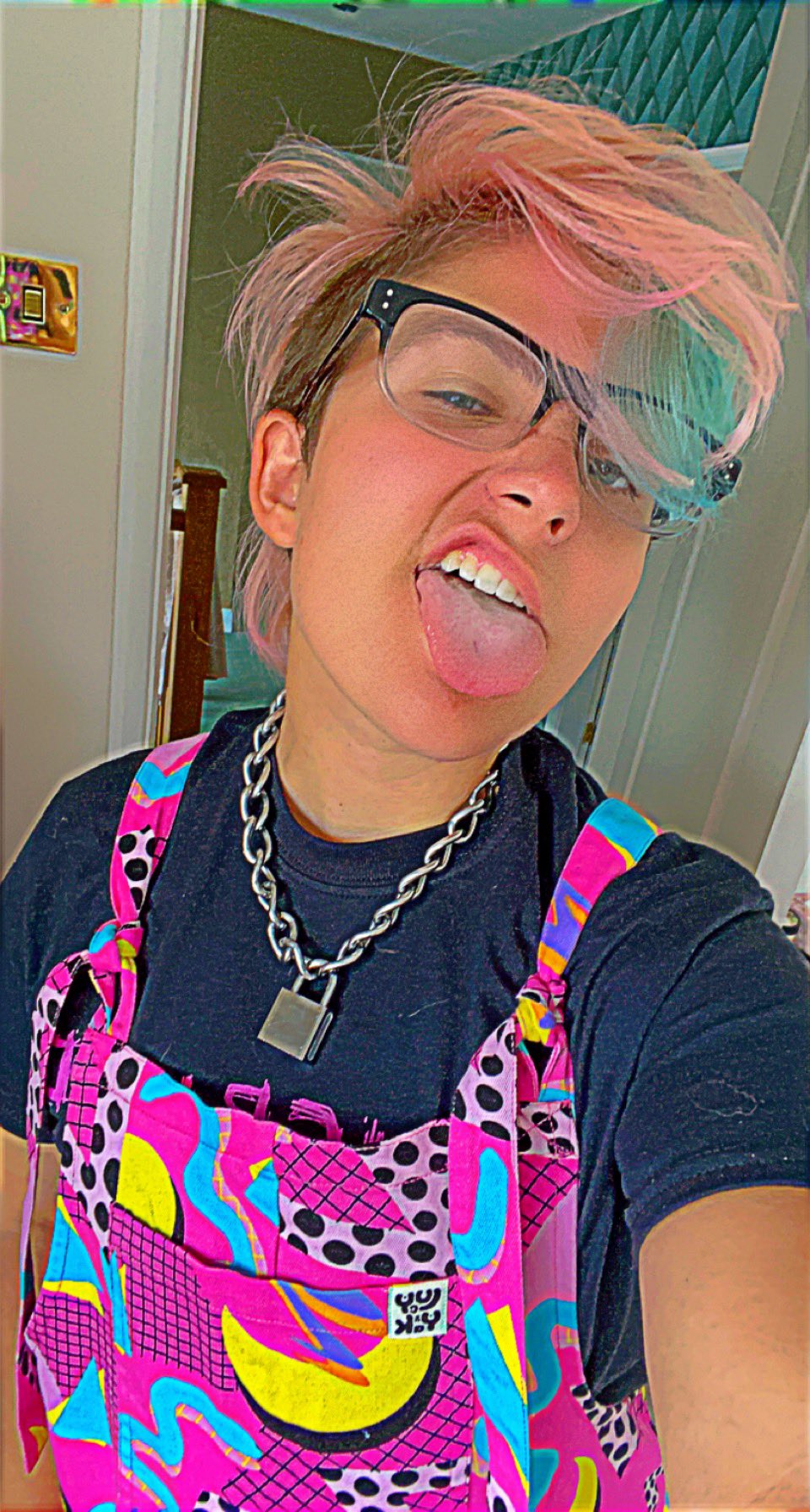Non-binary Yungblud fan Wolfie pictured wearing pink dungarees with their tongue out. They are wearing glasses and have pink hair and a padlock around their neck.