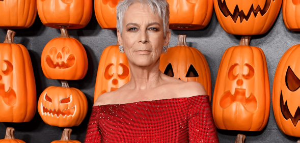 Jamie Lee Curtis wears a red outfit as she stands in front of a wall filled with orange pumpkin items shaped to look like jack-o-lanterns