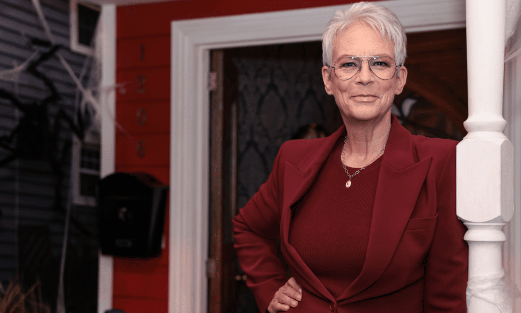 Jamie Lee Curtis wears a red outfit as she leans against the railing of a house
