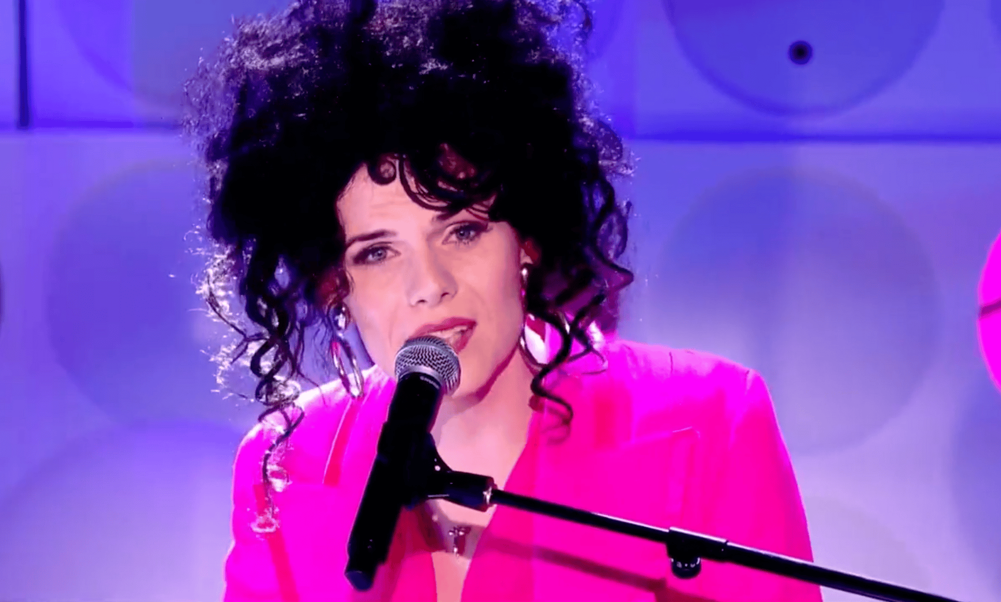 Jordan Gray wears a pink suit as she sings into a microphone during her Channel 4 Friday Night live performance