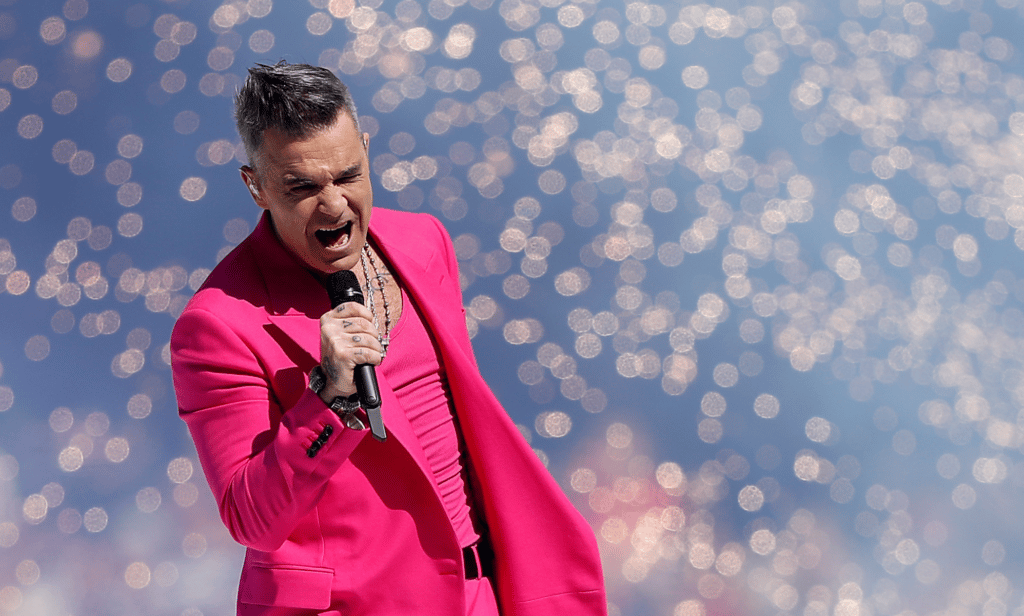 Robbie Williams wears a pink outfit as he sings into a microphone