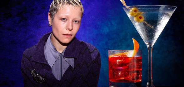 A photo of actor Emma D'Arcy sitting next to two drinks that are placed on a table