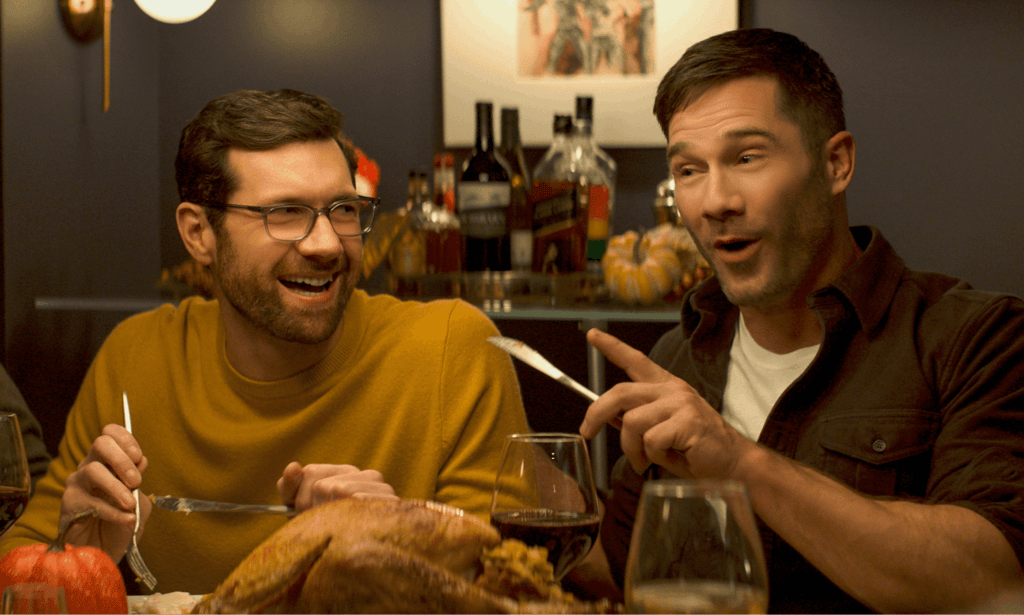 Billy Eichner and Luke Macfarlane at a dinner table, laughing with cutlery raised mid-gesture, in Bros