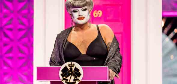 Drag Race UK queen Danny Beard standing at a pink podium in front of a pink front door