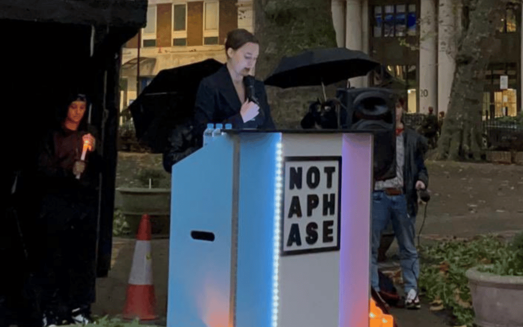 A screenshot from Twitter shows Kate Litman speaking at a podium during the Soho Trans Day of Remembrance.