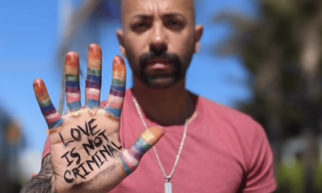 Dr Nas Mohamed, only publicly out LGBTQ+ Qatari, stands with his hand in front of him. His palm is facing towards the camera, and there are painted stripes in the colours of the LGBTQ+ and trans Pride flags on his fingers. In the centre of his palm, there are the worlds 'Love is not criminal'