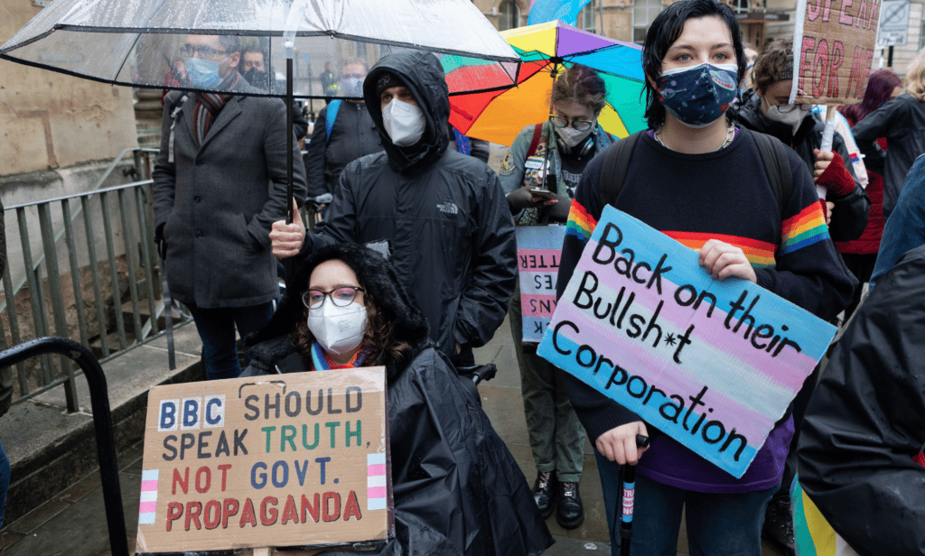 People hold up signs condeming an anti-trans article published by the BBC. One sign reads 'Back on the bullshit corporation' while the other reads 'bbc should speak truth not gov't propaganda'