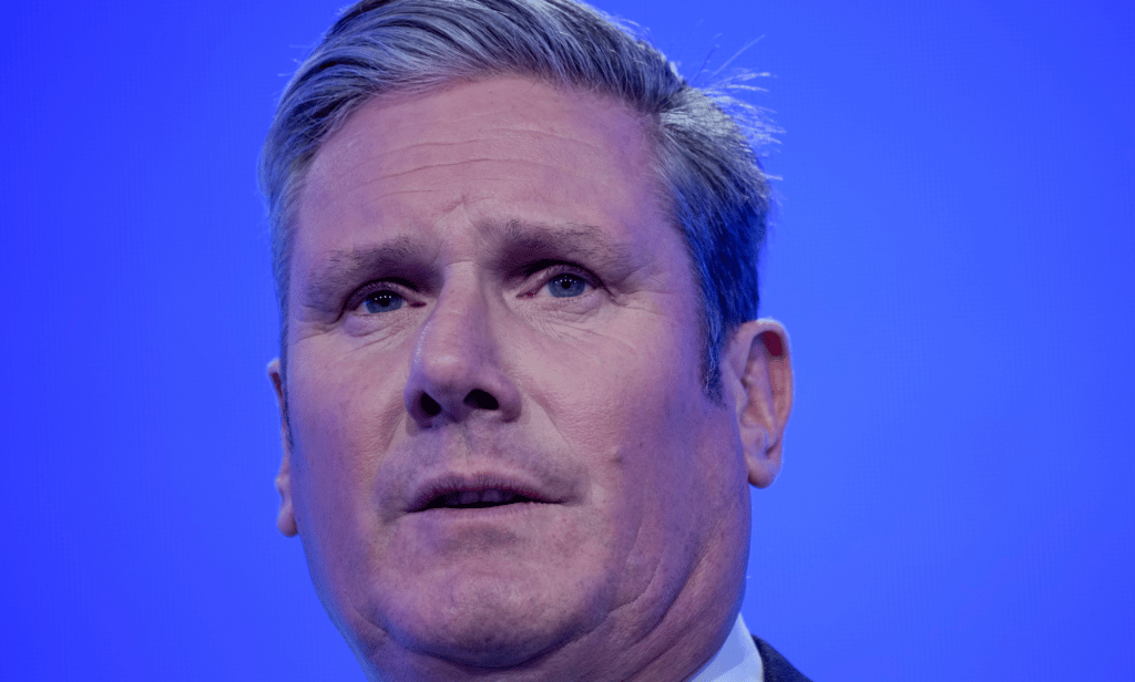 Labour Party leader Keir Starmer speaks to a crowd off camera as he stands in front of a blue background