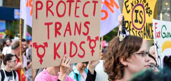 A photo shows a group of trans activists protesting, one sign being held says "Protect Trans Kids".