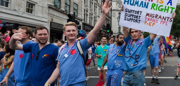 A group of NHS England doctors march during a Pride parade. One is wearing a sign that reads: "HEALTH IS A HUMAN RIGHT. Support Migrants, Support Trans PPL, Support PrEP"