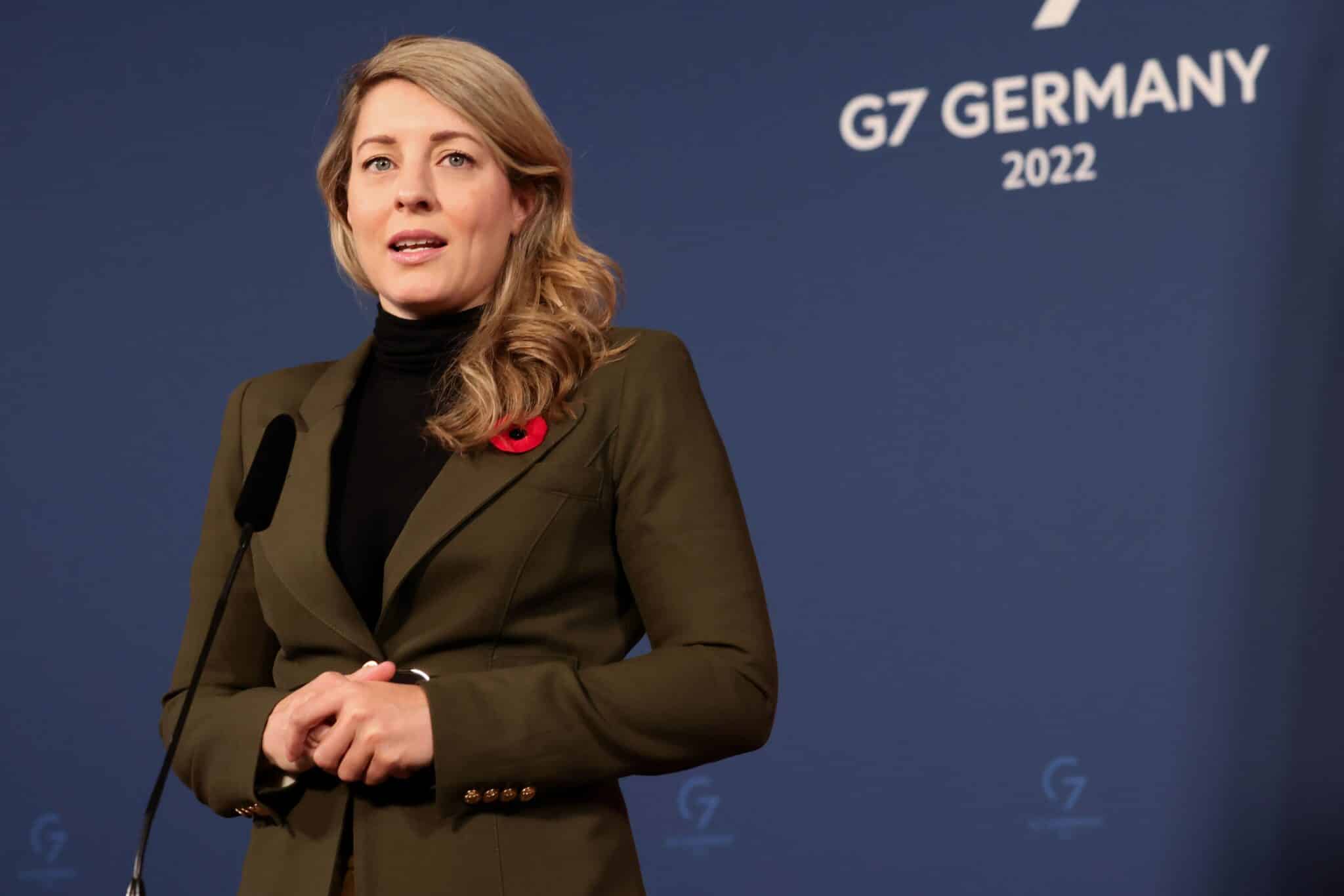 Melanie Joly speaks during the 67 infront of a navy blue backdrop, while wearing a dark green suit and a poppy.