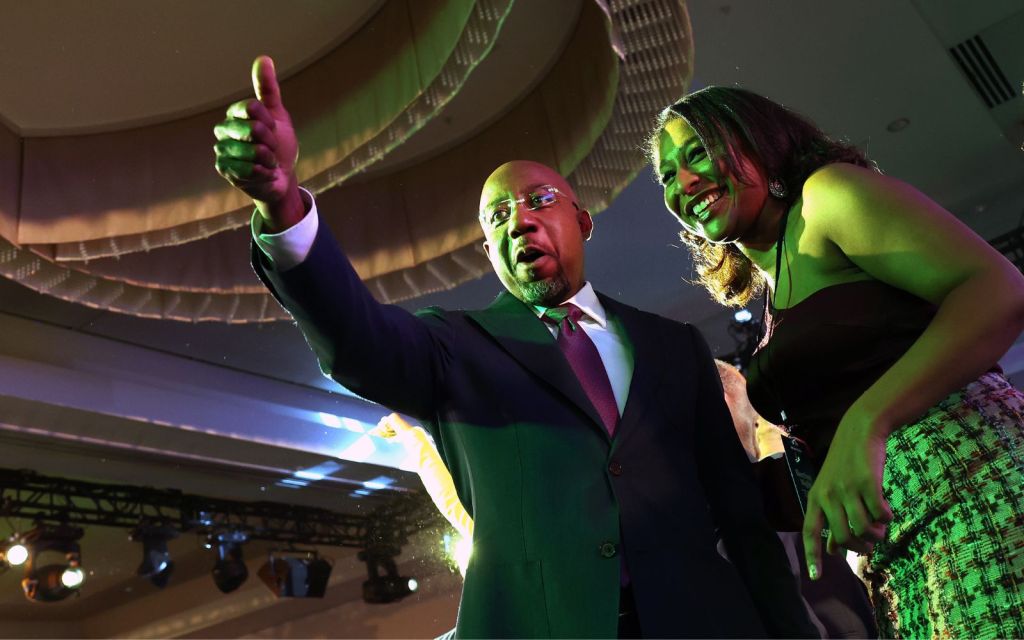Raphael Warnock gives a thumbs up while standing next to a person in a green light.