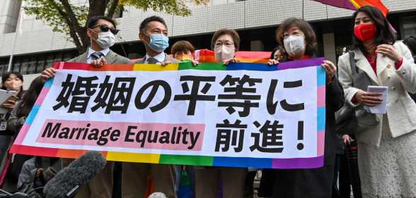 Plaintiffs holding up a rainbow-themed banner supporting marriage equality as a rainbow flag waves in the background.