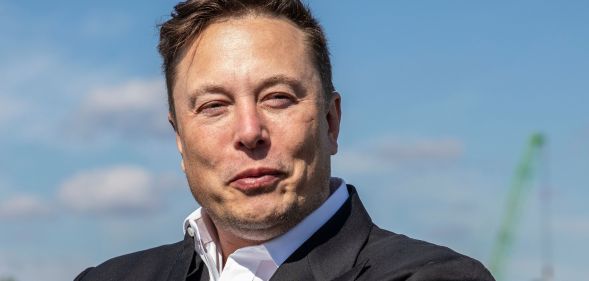 Elon Musk wears a white shirt and dark suit jacket as he stares somewhere off camera