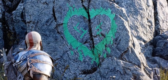 A screenshot of the game God of War Ragnarok, where the player looks at a green heart symbol painted on the side of a rock, containing Nordic symbols that translate to "SH" and "JS"