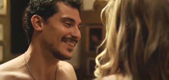 A still from The White Lotus showing actor Stefano Gianino as Niccolo smiling as he strips down in front of a woman with blonde hair who is turned away from the camera